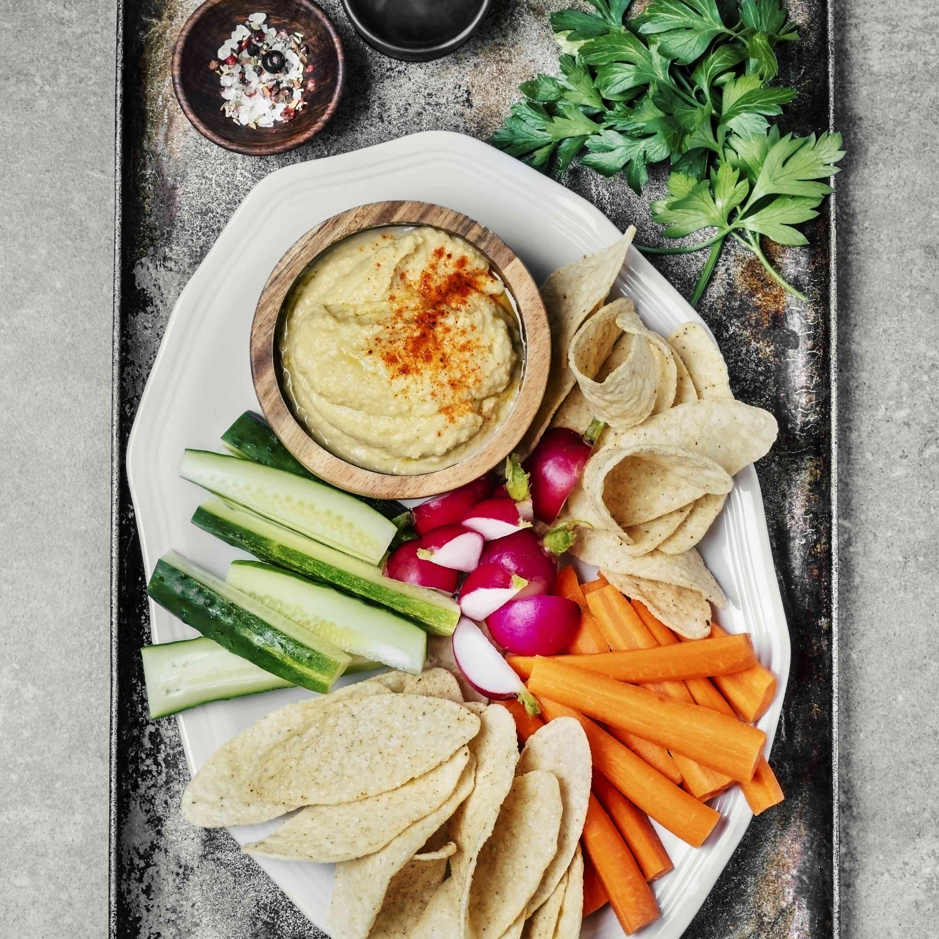 灰色背景上的金属托盘上有一盘蔬菜和一碗鹰嘴豆泥