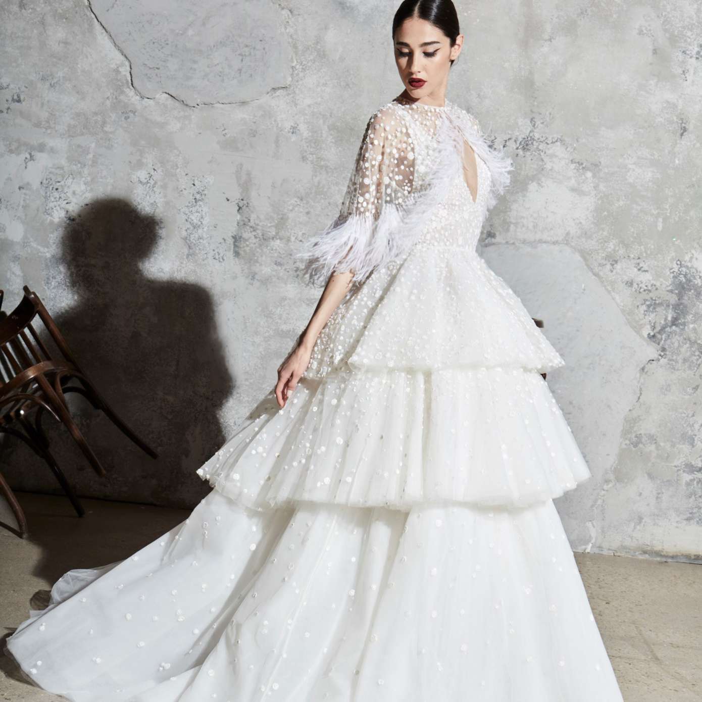 模特身穿带有亮片的v领舞会礼服和三层裙，搭配带有羽毛细节的薄纱耸肩