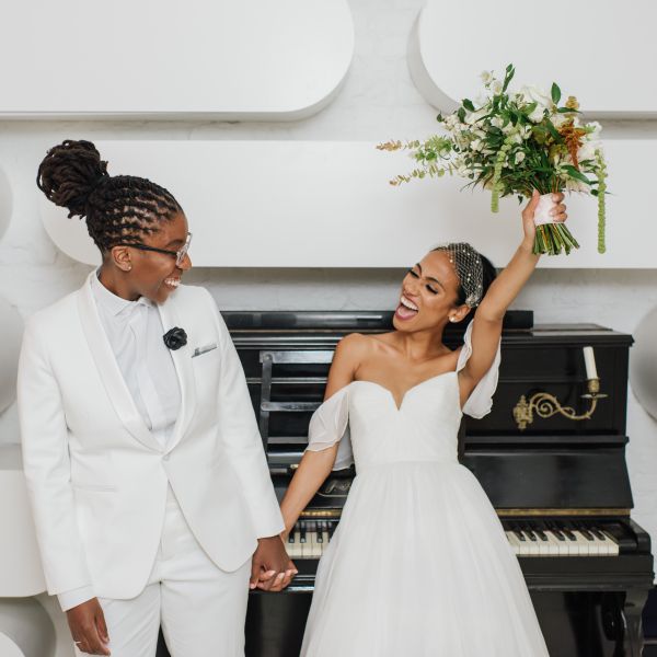 两位新娘手牵着手，在钢琴前摆姿势，左边的新娘手持花束，微笑着