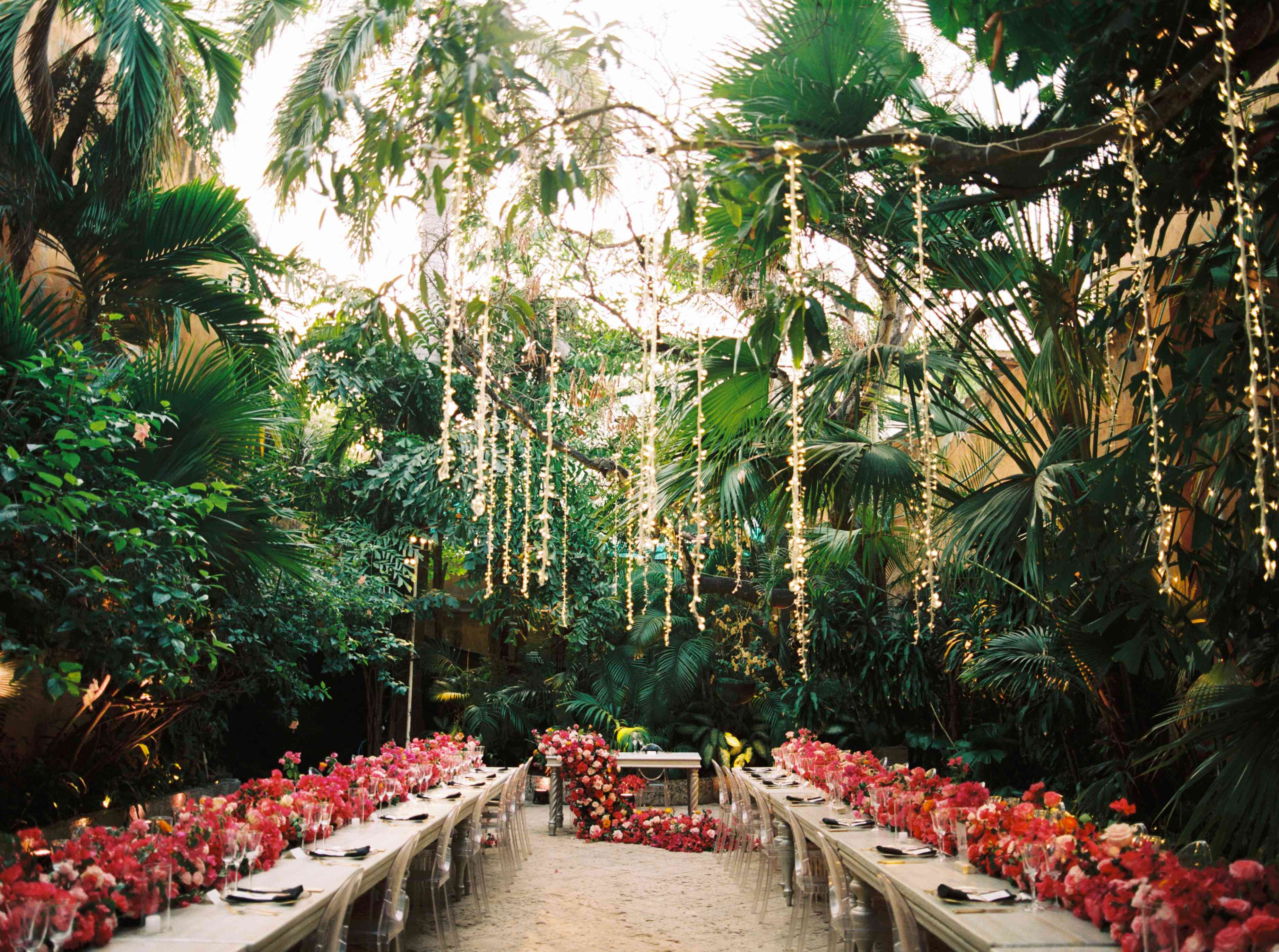 一串串的灯垂直地挂在热带树上，上方的长桌上排列着粉红色的花束