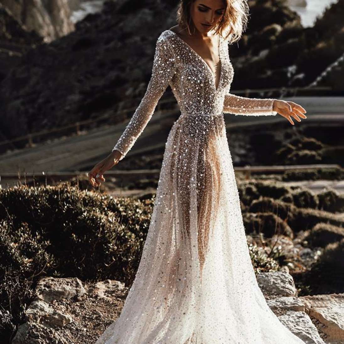Bride in long-sleeve silver gown standing on rocky cliffs above winding road and water
